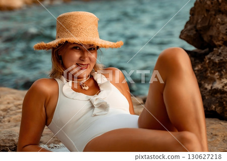Woman Beach Sunbathing Summer - A woman in a white swimsuit relaxes on a rocky beach while wearing a straw hat, enjoying the summer sun. Woman Beach Sunbathing Summer - A woman in a white swimsuit relaxes on a rocky beach while wearing a straw hat, enjoying the summer sun. 130627218