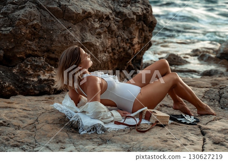 Woman Beach Relaxation - A woman in a white bikini and sunglasses sits on the beach with her legs crossed, looking out at the ocean. 130627219