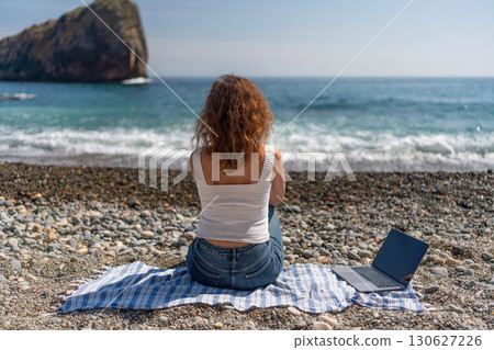 Beach Laptop Woman: Back view of woman using laptop on pebble beach, enjoying coastal views in daytime. 130627226