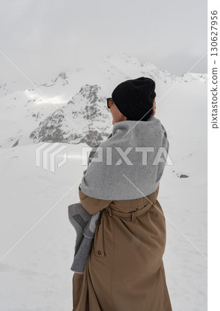 A woman wrapped in a scarf stands with her back to the camera, facing snowy mountains under a cloudy sky. Female solo travel, winter well-being A woman wrapped in a scarf stands with her back to the camera, facing snowy mountains under a cloudy sky. Female solo travel, winter well-being 130627956