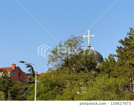 Church dome with metal cross above trees against blue sky. Religious symbol, cultural heritage and community identity with spirituality, belief and sacred landmark. 130627973