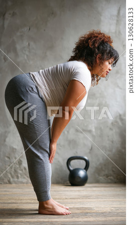 Plus-size Middle Eastern woman bending forward in standing yoga pose with kettlebell in background Plus-size Middle Eastern woman bending forward in standing yoga pose with kettlebell in background 130628133