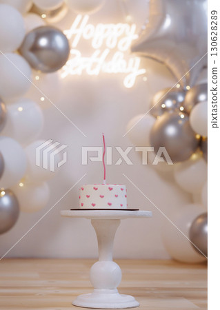 Birthday cake with one tall candle on white stand, balloons and glowing lights background Birthday cake with one tall candle on white stand, balloons and glowing lights background 130628289