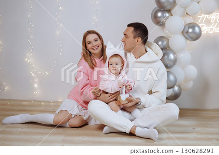 Happy young family with baby celebrating birthday on wooden floor near white wall and balloons 130628291