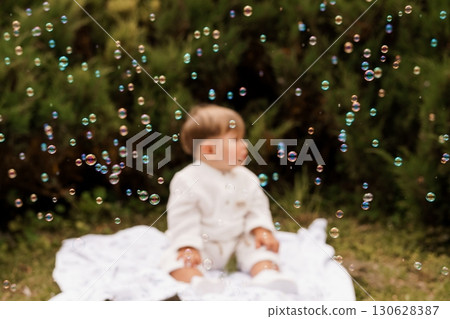 Adorable Baby Boy Playing with Bubbles Outdoors: Soft Focus, Green Garden, Caucasian Child, White Clothing, Joyful Moment, Natural Light Photography. 130628387