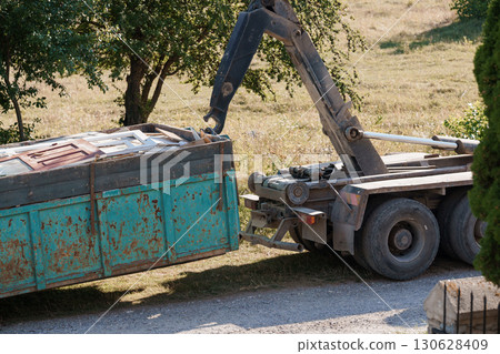 Rusty Dumpster Being Lifted by Crane Truck on Asphalt Roadside with Green Field Backdrop Rusty Dumpster Being Lifted by Crane Truck on Asphalt Roadside with Green Field Backdrop 130628409