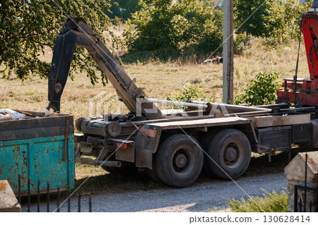 Construction Debris Removal with Crane Truck in Field: Rusty Container, Gray Road, Industrial Vehicle, Green Foliage, Sunny Day 130628414