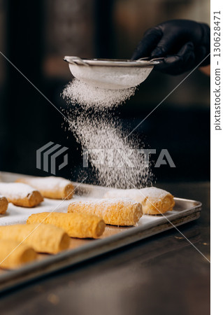 Chef dusting pastries with powdered sugar on tray in modern bakery kitchen 130628471