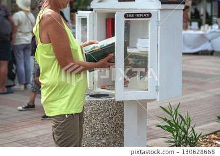 Woman taking book from outdoor street library cabinet with sign Take and Replace. Concept of free reading, knowledge sharing, urban community culture 130628668