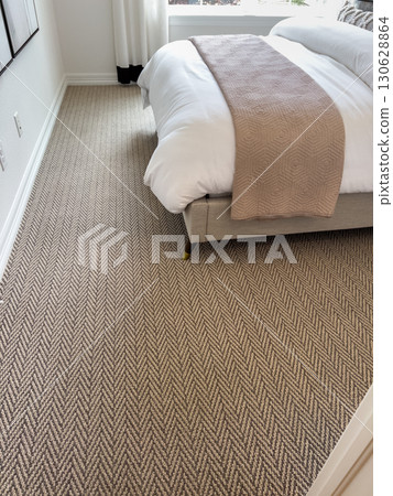 A side view of a modern bed with white linens and a beige blanket shows natural light pouring in through a nearby window. The room features a textured area rug. 130628864
