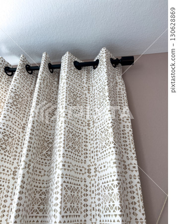 Detailed vertical shot of white and beige patterned curtains with grommets on a black curtain rod in a bright room. 130628869