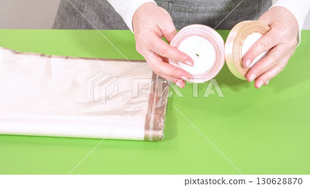Hands hold a roll of gold ribbon and satin-edged tissue paper over a bright green table. These materials are used to wrap a floral bouquet. 130628870