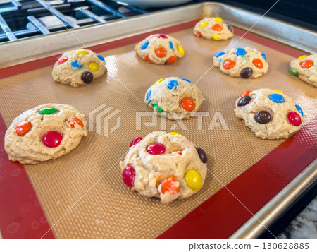 Round cookie dough balls with chocolate candies are placed on a silicone baking mat inside an oven tray. The unbaked cookies are arranged neatly and brightly lit. 130628885