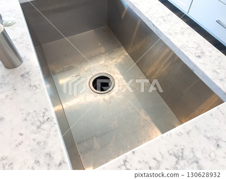 A clean stainless kitchen sink is set into a light marble-style countertop. The photo emphasizes a minimal, polished kitchen layout from above. 130628932