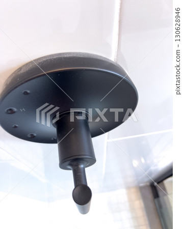 Close-up view of a matte black wall-mounted shower head in a modern tiled bathroom. The fixture is minimalist in style and set against clean white tiles with reflections of natural light. Close-up view of a matte black wall-mounted shower head in a modern tiled bathroom. The fixture is minimalist in style and set against clean white tiles with reflections of natural light. 130628946