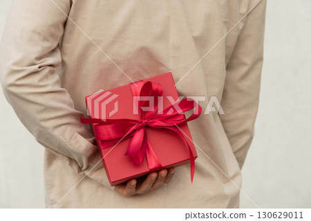 Man hiding red gift box tied with ribbon behind their back, wearing beige shirt for surprise girlfriend. Birthday present suggests surprise or special occasion. Valentine's Day. 130629011