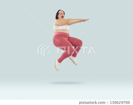 Fat funny excited woman wearing sportswear jumping with closed eyes on studio blue background. 130629700