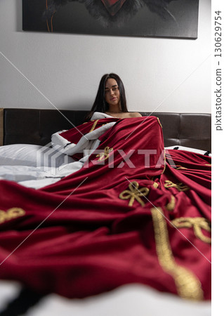 girl sits comfortably in bed draped in a rich royal robe adorned with golden details. The relaxing atmosphere of the room enhances the serene moment of leisure. girl sits comfortably in bed draped in a rich royal robe adorned with golden details. The relaxing atmosphere of the room enhances the serene moment of leisure. 130629754