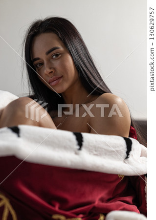 girl with long dark hair lounges on a bed comfortably wrapped in a rich royal robe. Her calm expression suggests relaxation and peace in a cozy atmosphere. 130629757
