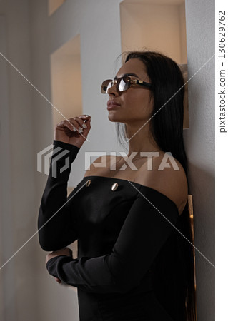 girl with glasses stands gracefully indoors showcasing an off shoulder black outfit. Soft lighting highlights her profile as she ponders thoughtfully. 130629762