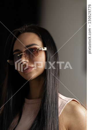 girl with long dark hair and trendy glasses poses confidently in a chic environment. The soft lighting highlights her features creating a captivating atmosphere around her. girl with long dark hair and trendy glasses poses confidently in a chic environment. The soft lighting highlights her features creating a captivating atmosphere around her. 130629776