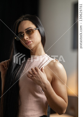 girl with long hair and glasses stands indoors exuding self assurance. She gently adjusts her clothing highlighting her strong physique. The setting has a modern feel with soft lighting. girl with long hair and glasses stands indoors exuding self assurance. She gently adjusts her clothing highlighting her strong physique. The setting has a modern feel with soft lighting. 130629781