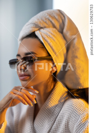 In a warm setting a girl with a towel on her head relaxes in her kitchen after a shower. Dawn light shines through the window as she enjoys a moment of calm while wearing glasses. 130629783