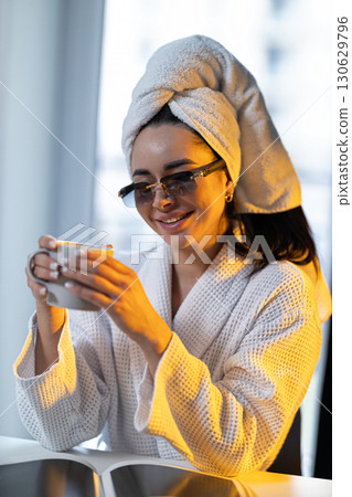 young girl sits comfortably in bright kitchen after her shower wearing glasses and towel on her head savoring warm drink dawn light fills the room.. 130629796