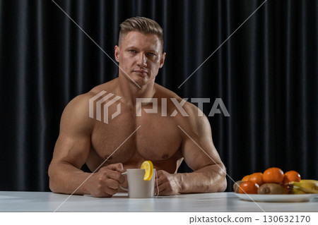 athletic man relaxes at table in cozy indoor space holding warm mug tea garnished with lemon slice surrounded by a colorful assortment of fresh fruit. 130632170