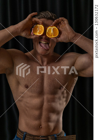 muscular individual is playfully holding orange halves in front of his eyes while sticking out his tongue. The setting is a studio with a dark background highlighting his physique. 130632172
