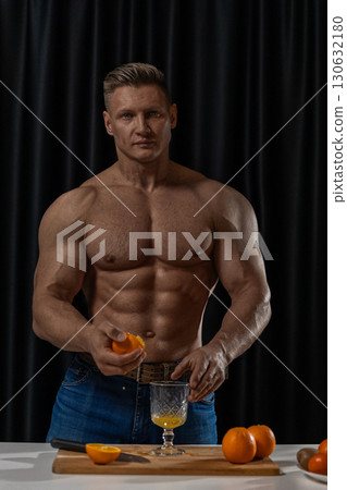 In a kitchen with dark curtains a muscular man is squeezing oranges into a glass while surrounded by fresh oranges. The focus is on his strength and commitment to healthy living. 130632180