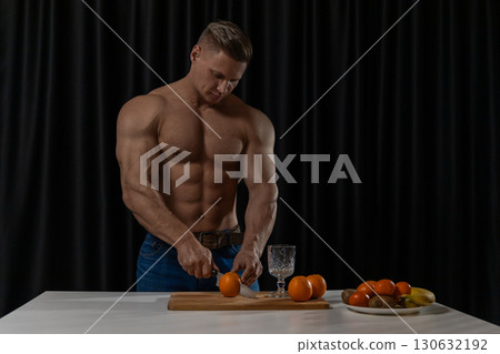 In a contemporary kitchen a muscular man is focused on cutting oranges. He stands at a white countertop preparing nutritious food. A bowl of fruits is nearby highlighting healthy eating. 130632192
