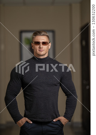 athletic man with glasses stands confidently in a modern indoor setting. His muscular build is emphasized by a fitted black shirt. The environment exhibits clean lines and neutral tones. 130632200