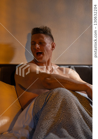 athletic man sits up in bed stretching and yawning as sunlight filters through the window. He relaxes on crisp sheets showcasing his toned physique in a cozy morning setting. athletic man sits up in bed stretching and yawning as sunlight filters through the window. He relaxes on crisp sheets showcasing his toned physique in a cozy morning setting. 130632204