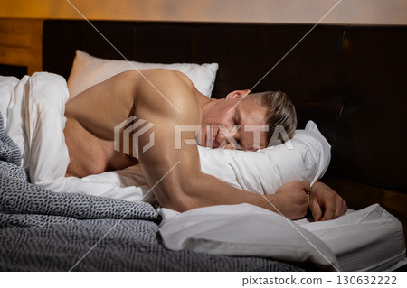 athletic man rests soundly in bed his muscular build relaxed against soft pillows and blankets. The warm lighting and cozy atmosphere indicate a calm end to a strenuous day. 130632222