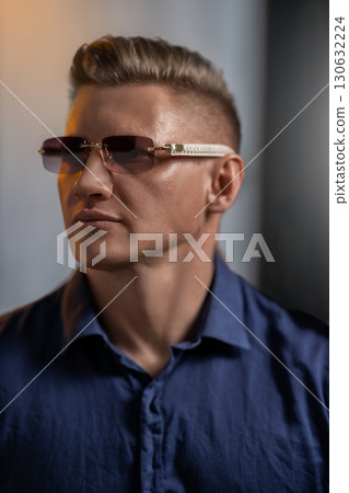 man with short hair and glasses poses confidently wearing a blue shirt. The warm evening light highlights his features creating a dramatic effect in the background. 130632224