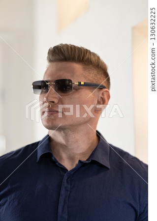 A man with short hair and stylish glasses gazes thoughtfully in a bright indoor environment wearing a dark blue shirt. The soft lighting enhances his features. A man with short hair and stylish glasses gazes thoughtfully in a bright indoor environment wearing a dark blue shirt. The soft lighting enhances his features. 130632225