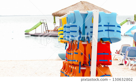 Colorful life jackets are neatly stacked on a beach, with a water slide and lounge chairs visible in the background, creating a vibrant summer scene Colorful life jackets are neatly stacked on a beach, with a water slide and lounge chairs visible in the background, creating a vibrant summer scene 130632802