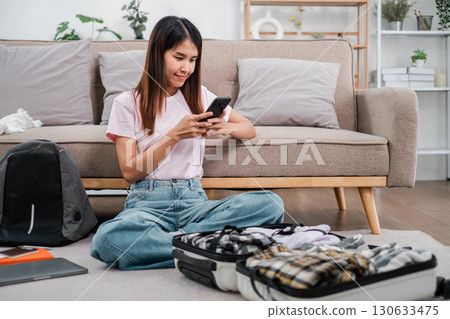 A young woman sits on the floor packing a suitcase while using her smartphone, preparing for a trip in a cozy living room. 130633475