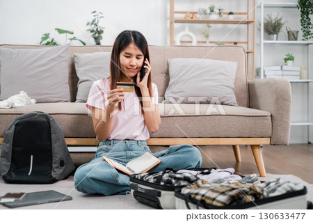 Woman sitting on floor packing suitcase, using phone and credit card, preparing for travel in a cozy living room. Woman sitting on floor packing suitcase, using phone and credit card, preparing for travel in a cozy living room. 130633477