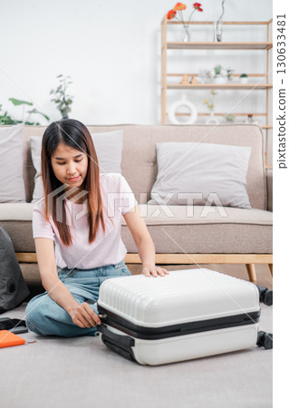 A young woman sits on the floor packing a suitcase in a stylish living room, preparing for a travel adventure or vacation. A young woman sits on the floor packing a suitcase in a stylish living room, preparing for a travel adventure or vacation. 130633481