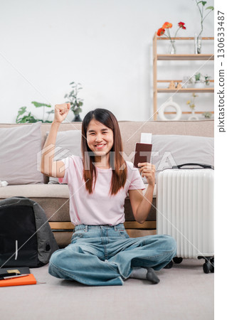 Smiling woman sitting on floor with passport and luggage, excited for travel. Cozy living room setting. 130633487