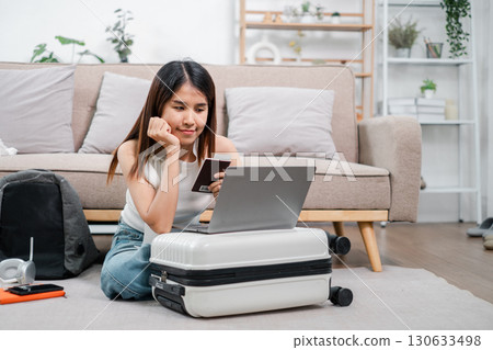 Woman using laptop and credit card for online shopping, sitting on suitcase in a modern living room. Woman using laptop and credit card for online shopping, sitting on suitcase in a modern living room. 130633498