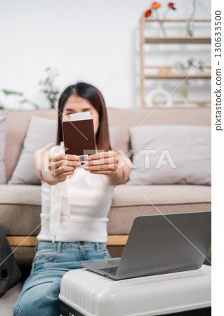 Woman sitting on floor holding passport, with laptop and suitcase, ready for travel in a cozy living room setting. Woman sitting on floor holding passport, with laptop and suitcase, ready for travel in a cozy living room setting. 130633500