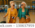 Two stylish elderly women at an airport, smiling with luggage, tickets, and a small dog in a handbag. Generated AI. 130637959