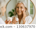 An elderly woman smiles while applying face cream in front of a round bathroom mirror. Generated AI. 130637970