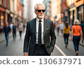 An older man in a sharp black suit and sunglasses confidently walks through a busy city street. Generated AI. 130637971
