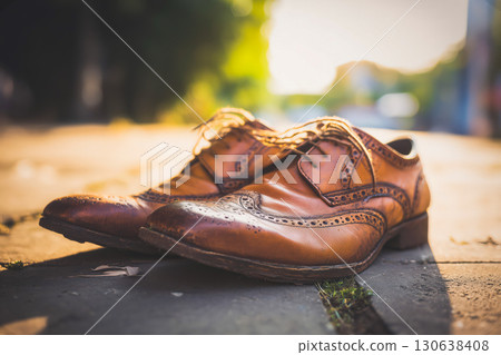 Pair of vintage brown leather brogues with decorative perforations resting on sunny pavement, worn leather showing scuffs and character, classic footwear in warm evening light scene 130638408