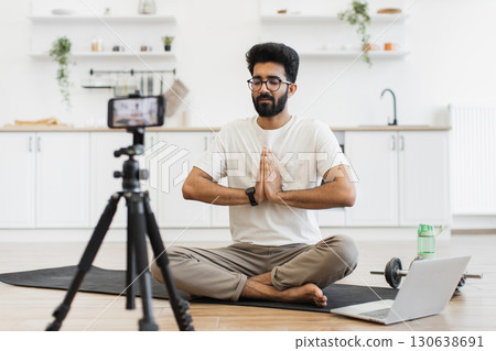 Young adult man sitting in modern kitchen recording video blog about fitness and mindfulness while meditating. Promoting healthy lifestyle including mental and physical wellness to his online audience Young adult man sitting in modern kitchen recording video blog about fitness and mindfulness while meditating. Promoting healthy lifestyle including mental and physical wellness to his online audience 130638691