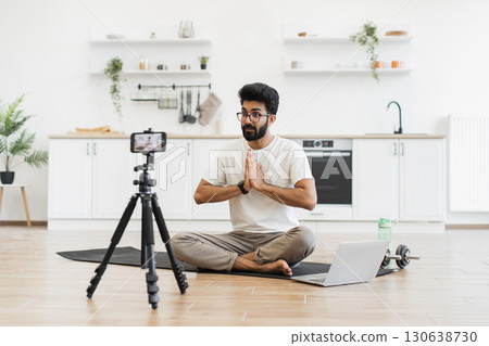 Young adult man sitting in modern kitchen recording video blog about fitness and mindfulness while meditating. Promoting healthy lifestyle including mental and physical wellness to his online audience 130638730